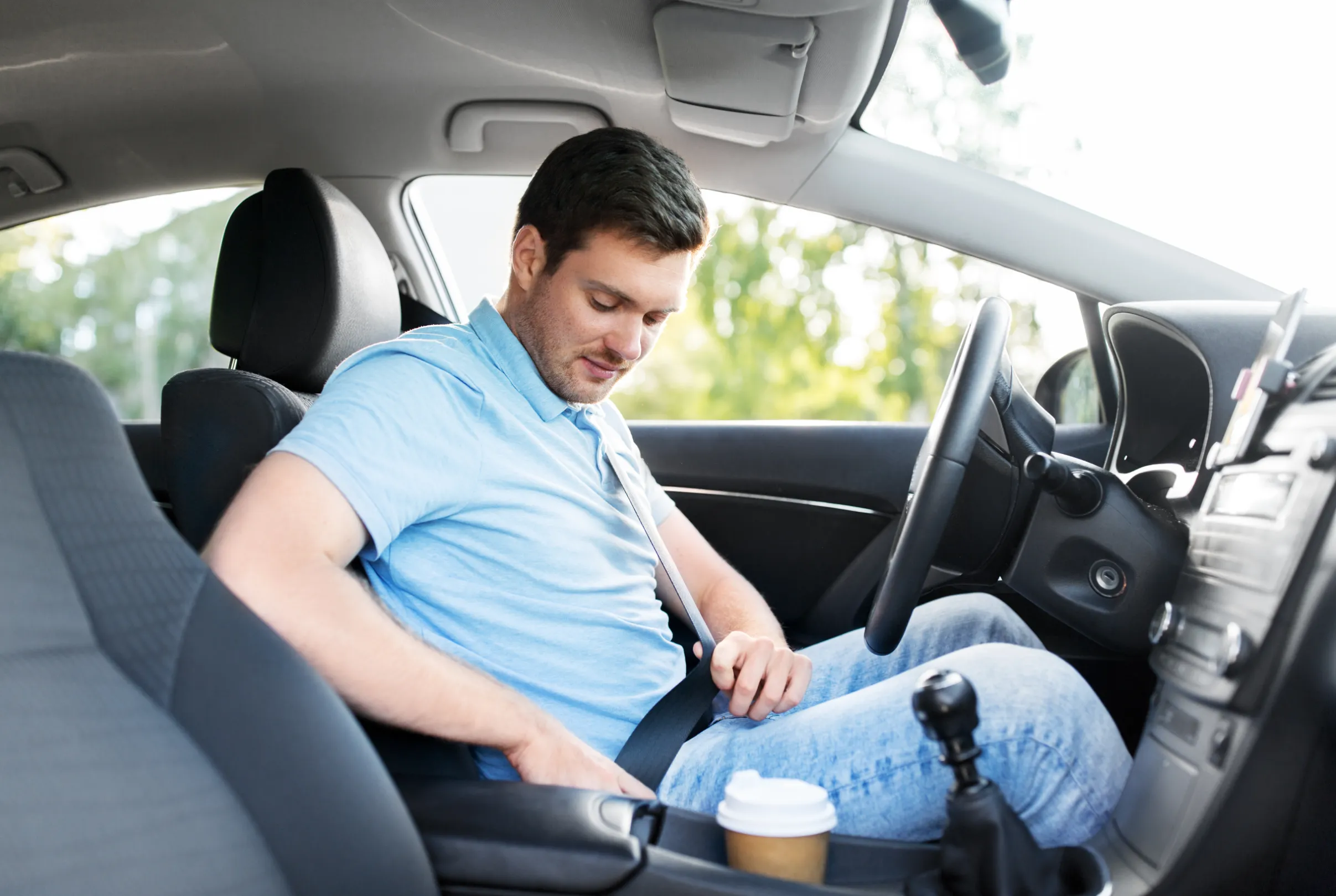 A driver seated in a car fastening a three-point seat belt, drawing the webbing down towards the buckle beside the seat.