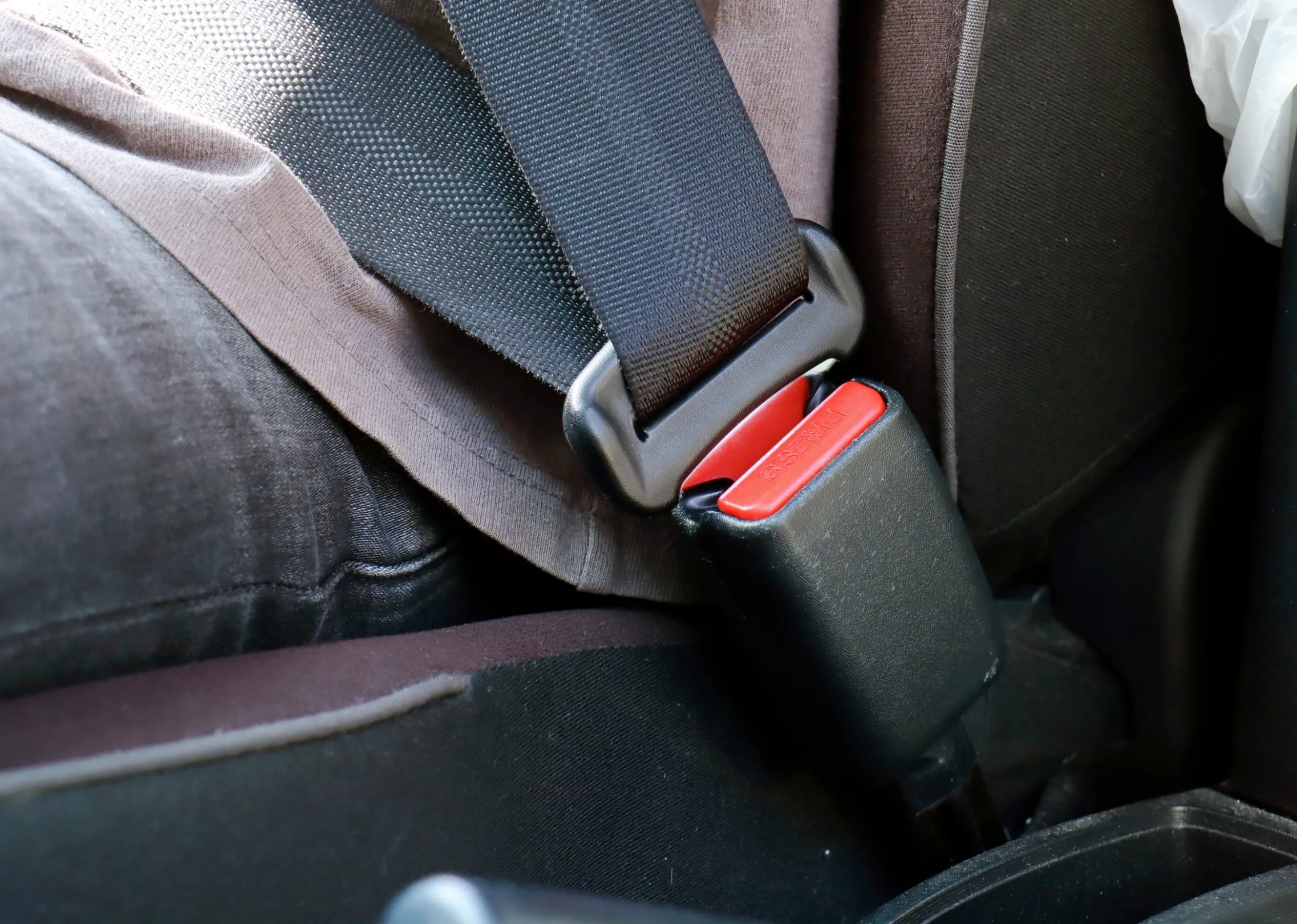 A three-point seat belt fastened across a seated occupant in a fabric-trimmed vehicle cabin, with the tongue plate engaged in the buckle.
