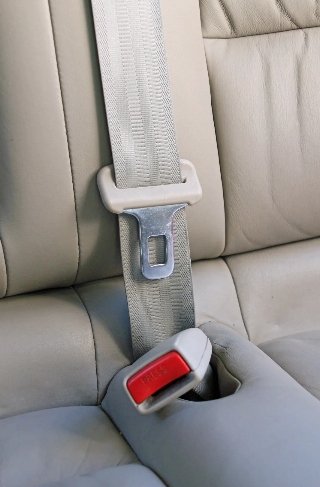 A three-point seat belt on a light-coloured leather rear seat, showing the shoulder webbing, tongue plate and buckle with a red PRESS release.
