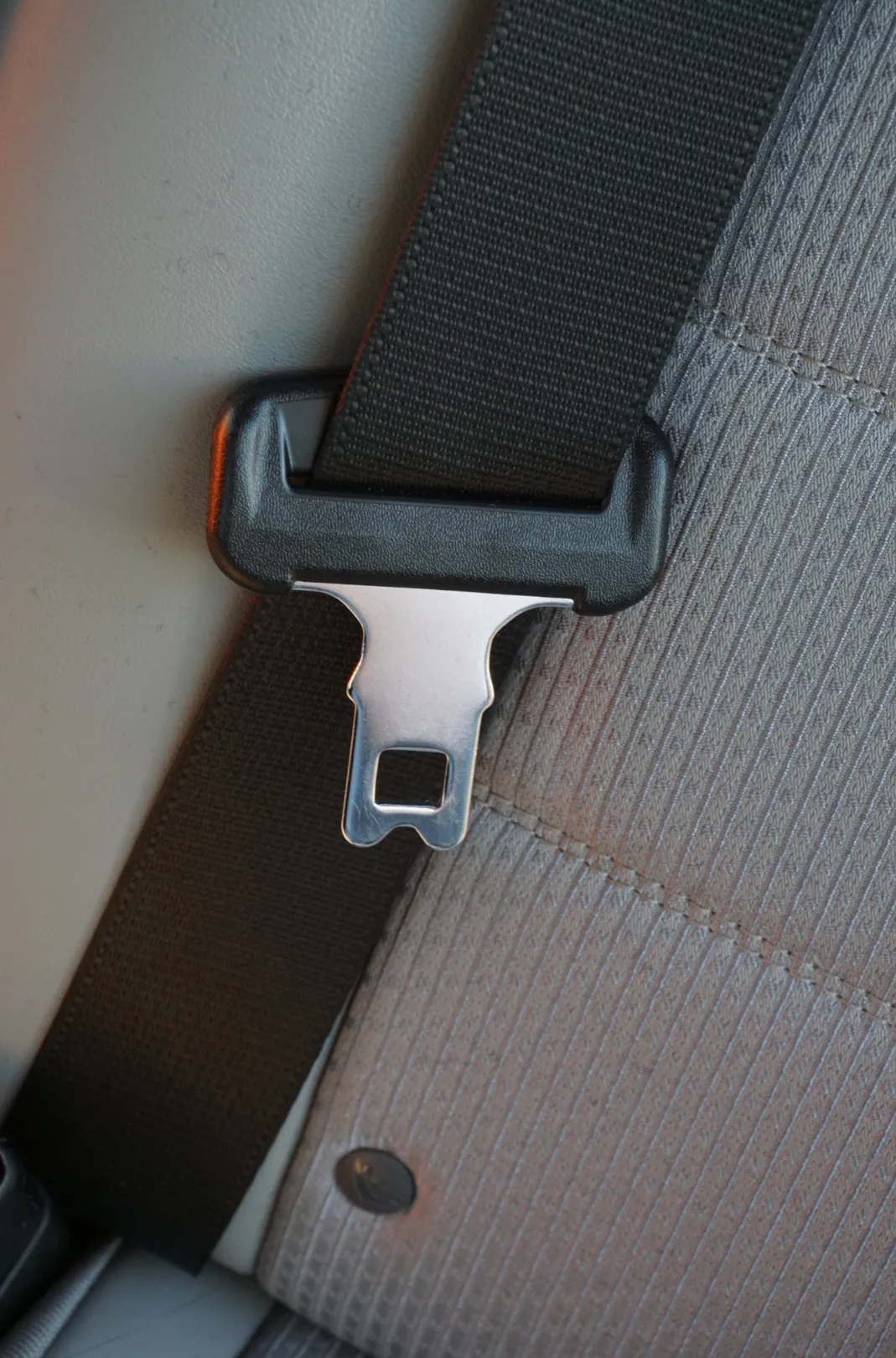 A polished steel seat belt tongue plate resting in a moulded webbing guide on a grey fabric vehicle seat.